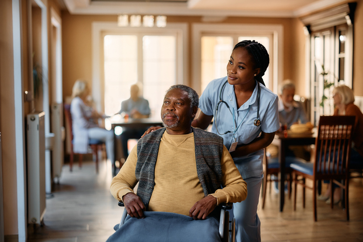 Nurse pushing man in wheelchair through dining area